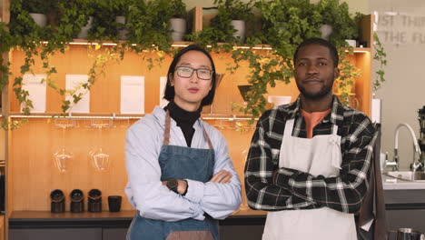 two multiethnic waiters with crossed arms smiling at camera while standing behind counter in a coffee shop 1