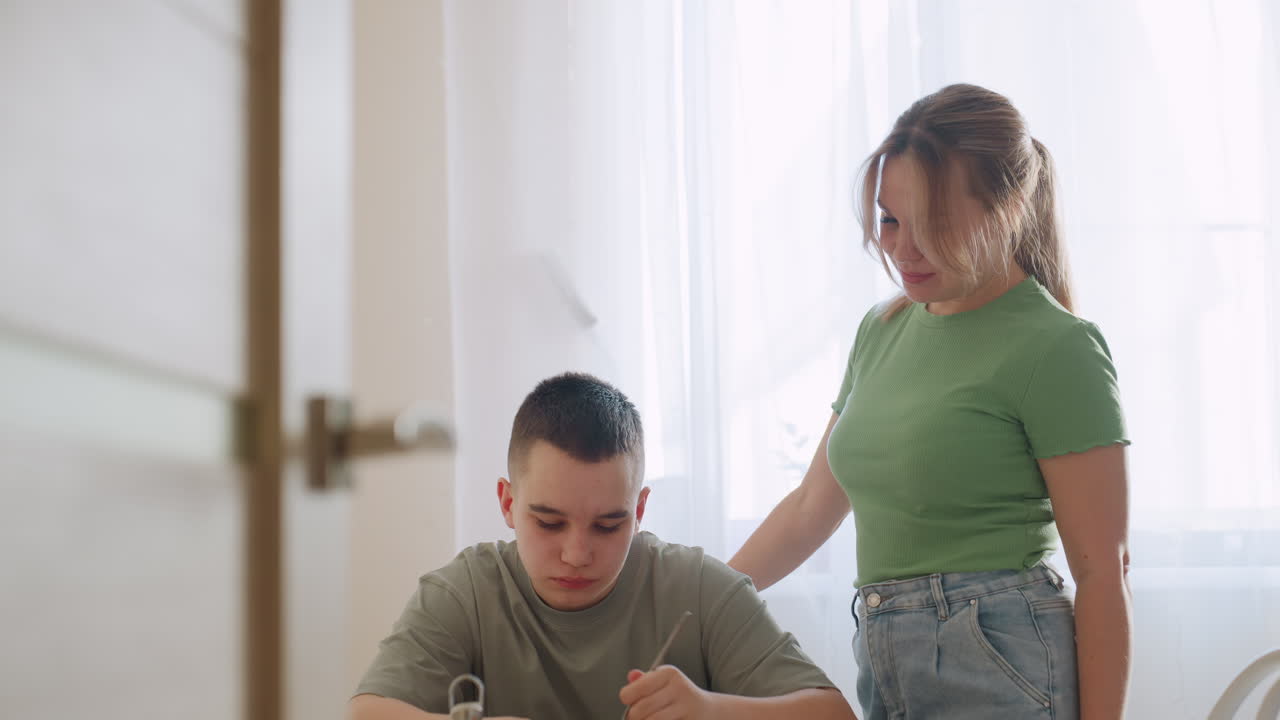 Loving mum stands close to son as he eats lunch with fork, smiling warmly while watching him enjoy fresh meal at home, natural daylight creating tender family moment of care