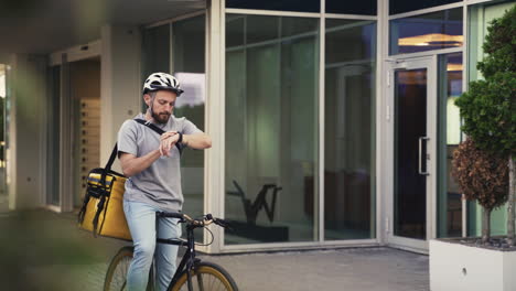 Food-Delivery-Guy-Wearing-Thermal-Backpack-Takes-A-Look-At-His-Smartwatch-To-Know-Where-To-Go-With-His-Bike