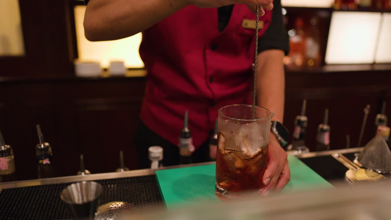 Bartender in red vest stirs whiskey cocktail with bar spoon under warm, ambient lighting
