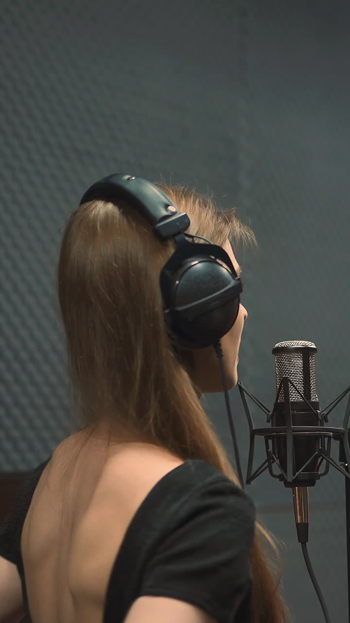 Positive woman vocalist with black headphones in dress with open back sings song to microphone dancing in recording studio close backside view
