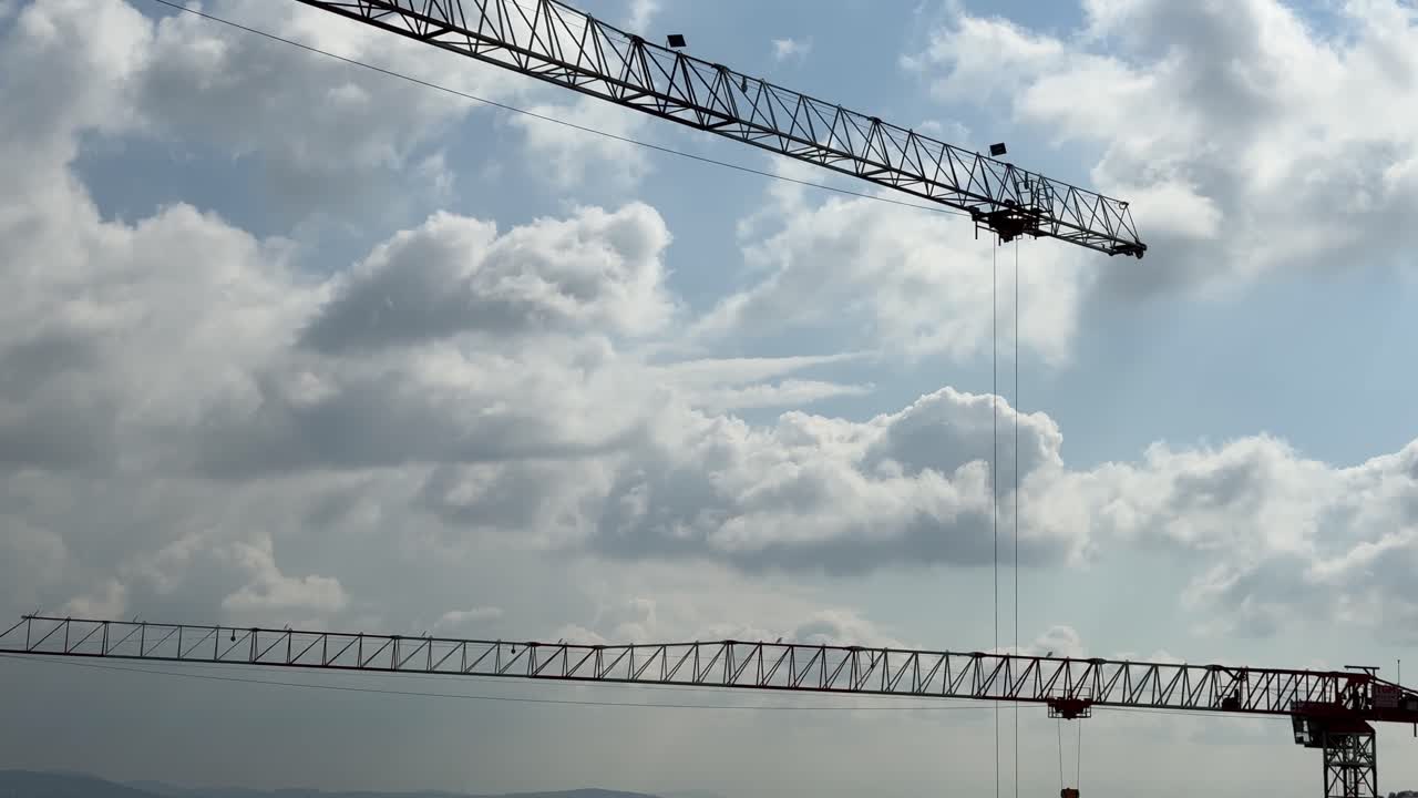 Low-angle shot showing the lattice structure (jibs) of two large tower cranes crossing against a partially cloudy, bright blue sky, indicating heavy urban construction or development