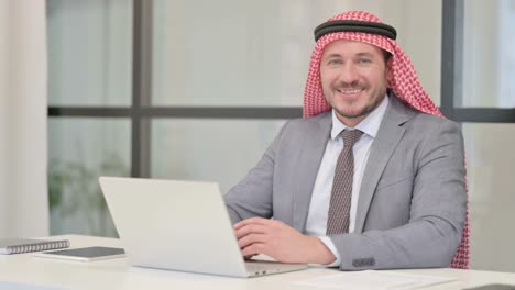 arab businessman smiling at camera while using laptop in office