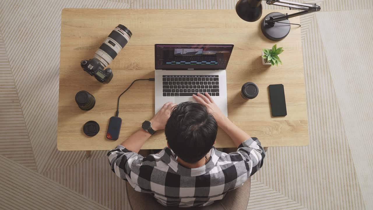 Top View Of A Male Editor Sitting In The Workspace Using A Laptop Next To The Camera Editing The Video At Home
