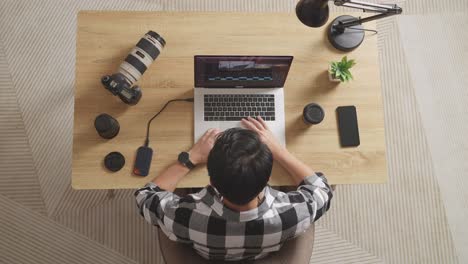 top view of a male editor sitting in the workspace using a laptop next to the camera editing the video at home