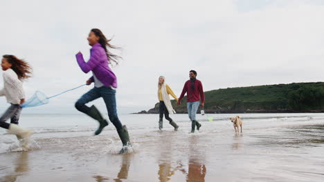 Multikulturelle-Familie-Mit-Hund,-Die-Im-Winterurlaub-Am-Strand-Entlang-Spaziert