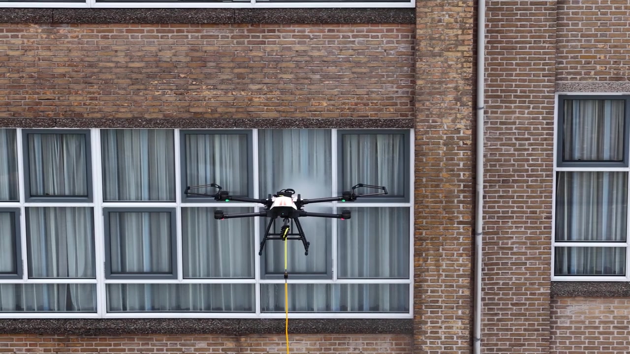 Drone autonomo con tubo e spruzzatore collegati che esegue un lavaggio ad alta pressione sulle finestre di un grande edificio in mattoni, rappresentando il futuro della tecnologia di pulizia industriale