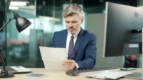 serious mature businessman in a formal suit examines a document sitting at work in office gray haired bearded focused business man reads a contract letter or documentation.