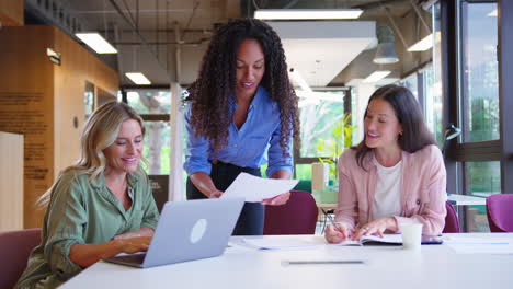 multi-cultural female business team sitting at desk with laptop in office collaborating on project