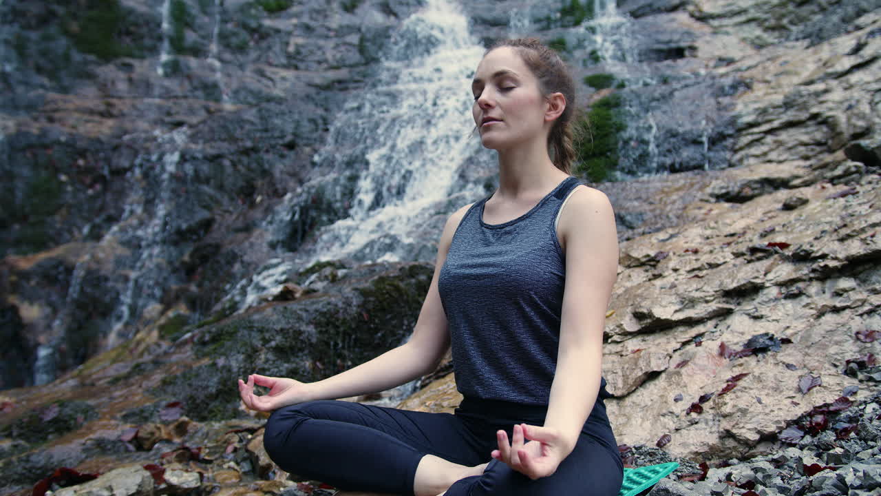 mujer practicando yoga, sentada en la postura de om en la roca cerca de una cascada, en la mano