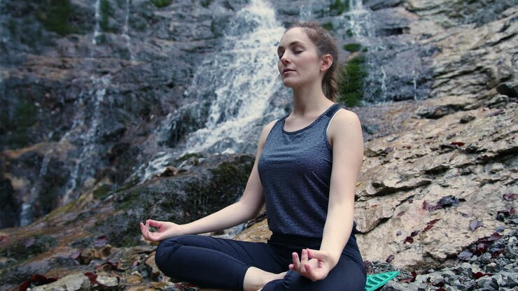 Mujer practicando yoga, sentada en postura Om sobre la roca cerca de una cascada, toma a mano alzada.