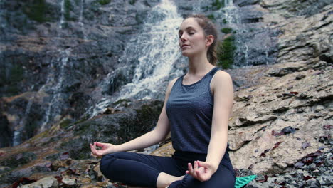 woman practicing yoga, sitting in om pose on the rock near a waterfall, handheld