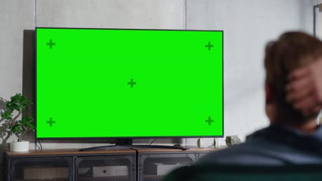 young handsome man sitting on a sofa and watching tv with horizontal green screen mock up. it's day time in room at home living room in loft apartment.