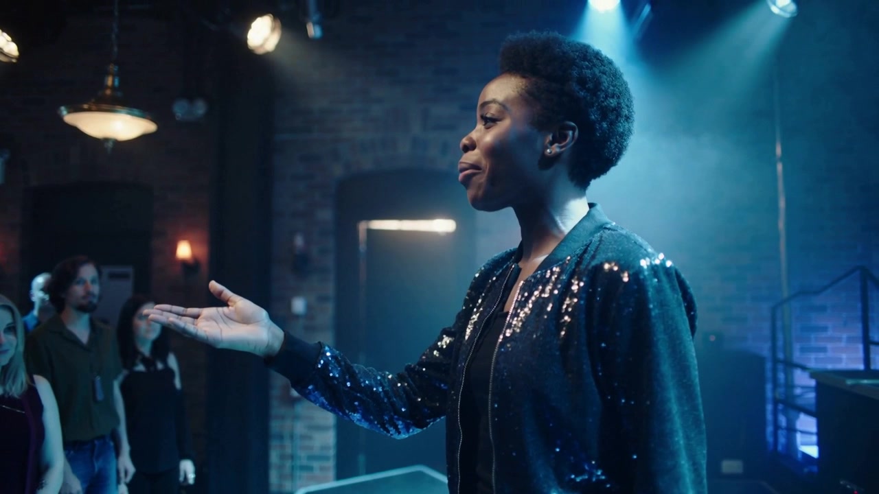 Luce blu illumina una cantante che si protende, coinvolgendo la folla su un palco di mattoni in un club, indossando una giacca di paillettes.