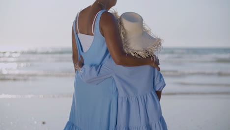 Back-View-Of-Mother-And-Daughter-Embracing-Each-Other-And-Watching-The-Sea-While-Standing-At-Seashore