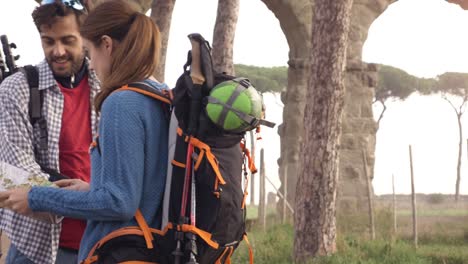 happy young couple backpackers tourists reading map guide for directions lost near roman aqueduct arches in parco degli acquedotti park ruins in rome at sunrise with guitar and sleeping bag slow motion steadycam