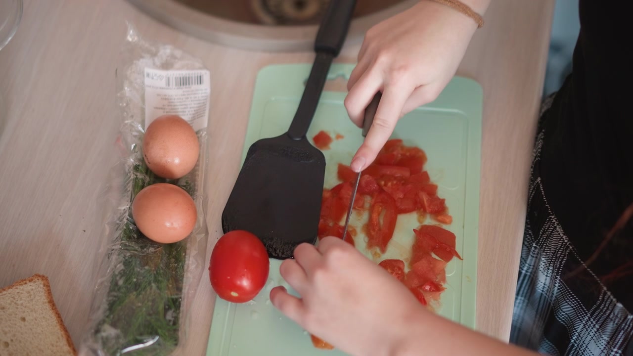 Donna bianca dai capelli rossi che affetta un pomodoro su un tagliere verde chiaro, spatola e uova allineate sul bancone, mani veloci e concentrate che preparano un pranzo rapido, cucina compatta di un appartamento, routine efficiente con un tocco giocoso.