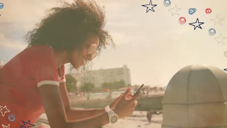colorful star icons against african american woman using smartphone near the beach