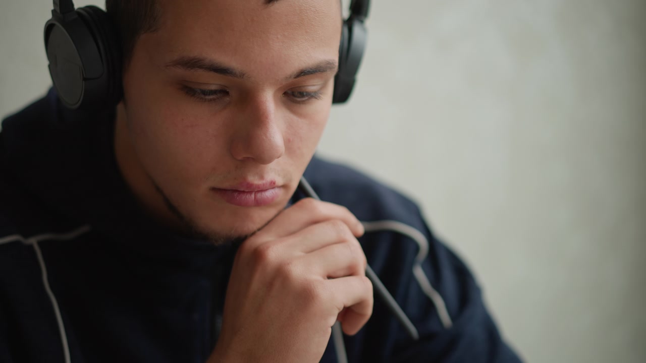 Gros plan sur un jeune homme portant un casque audio et posant son menton sur une main, tenant l'autre avec un stylo. Un éclairage doux met en valeur une expression calme sur un fond uni. Léger mouvement de caméra