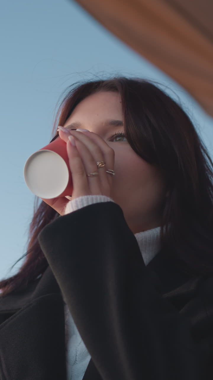 검은 코트를 입은 여성이 주스를 마시고, 앞에 노트북을 놓고, 컵을 떨어뜨리고, 계속 일하고, 배경에는 흐릿한 빨간 지붕과 장식 기둥이 있습니다.