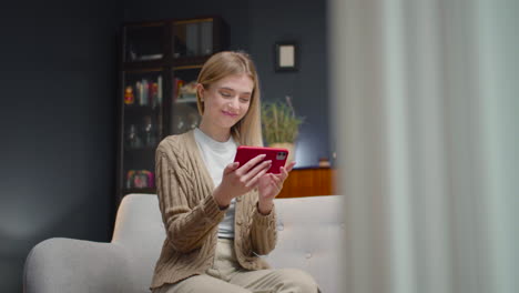 mujer joven feliz viendo videos divertidos en el teléfono inteligente y riendo mientras se sienta en un sofá en casa