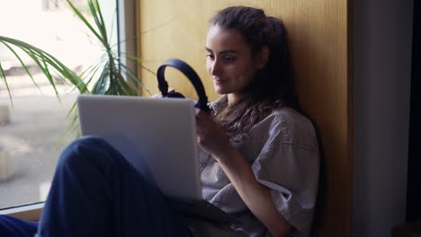 mujer freelancer sentada en el alféizar de la ventana en un café y trabajando en una computadora portátil con auriculares