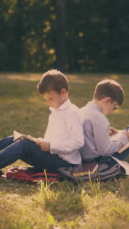 serious schoolmates sit back to back preparing for exams with textbooks on green lawn in garden wind turns pages