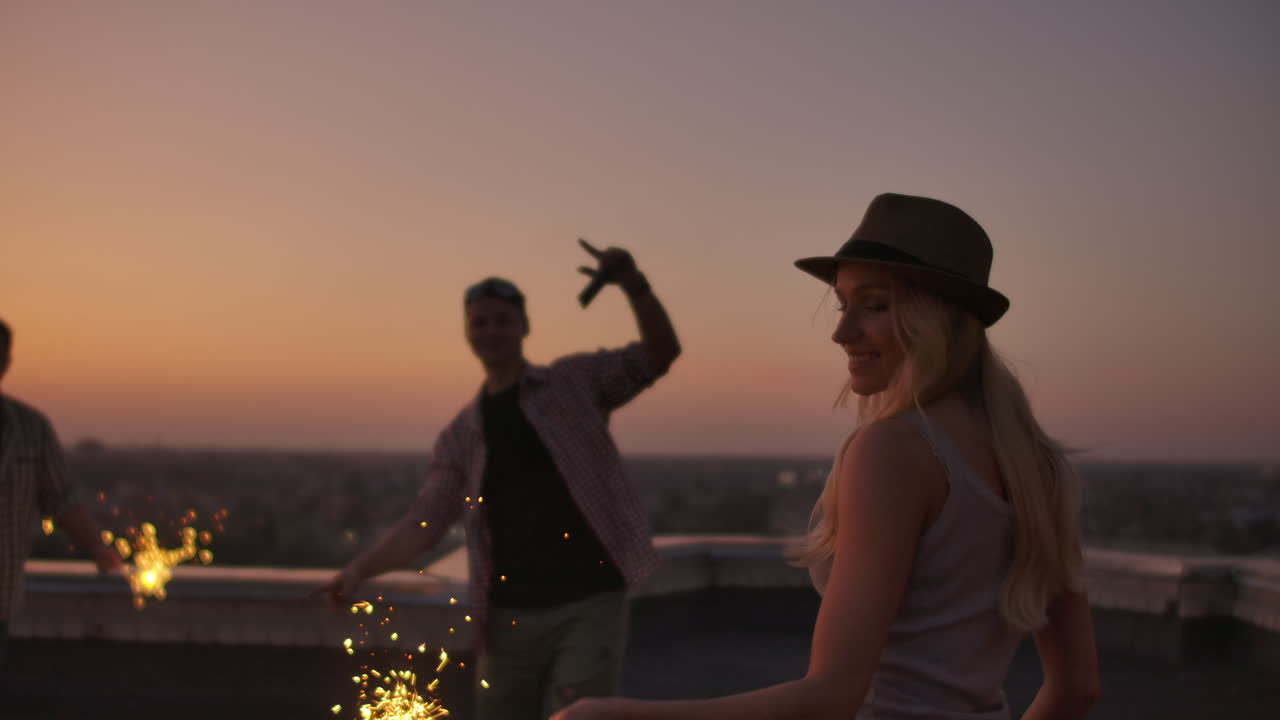 A company of girls and boys raise their hands up and jump on the roof with big bengal light in their hands. It's a crazy party at night.