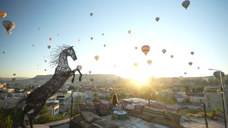 Einsame-Frau-Auf-Der-Terrasse-Mit-Blick-Auf-Heißluftballons,-Die-Bei-Sonnenaufgang-über-Der-Landschaft-Von-Kappadokien-In-Der-Türkei-Fliegen