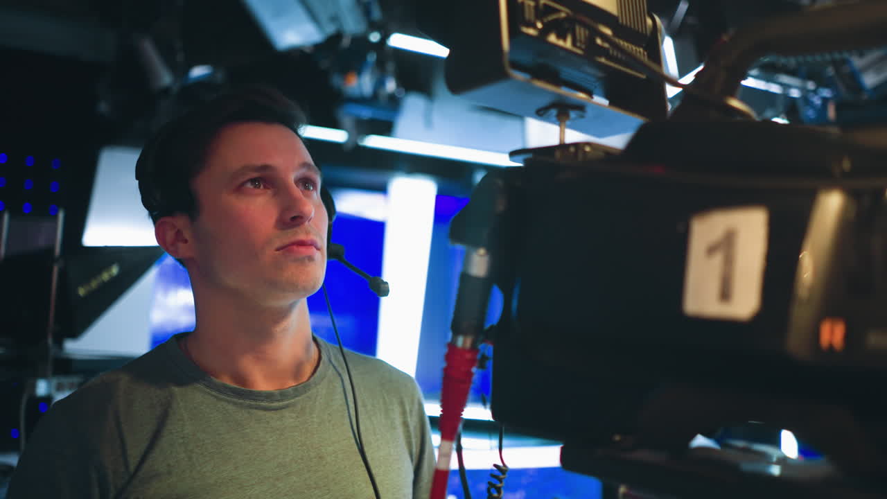 Focused male cameraman wearing headset operates broadcast camera inside brightly lit studio with blue background lights during live television production, concentrating on monitor while handling professional filming equipment