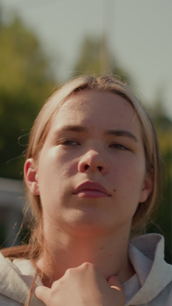 primer plano de una joven pensativa caminando en un estadio al aire libre con su mano apoyada en su sudadera con capucha, mirando pensativa, el fondo borroso presenta árboles verdes y postes eléctricos