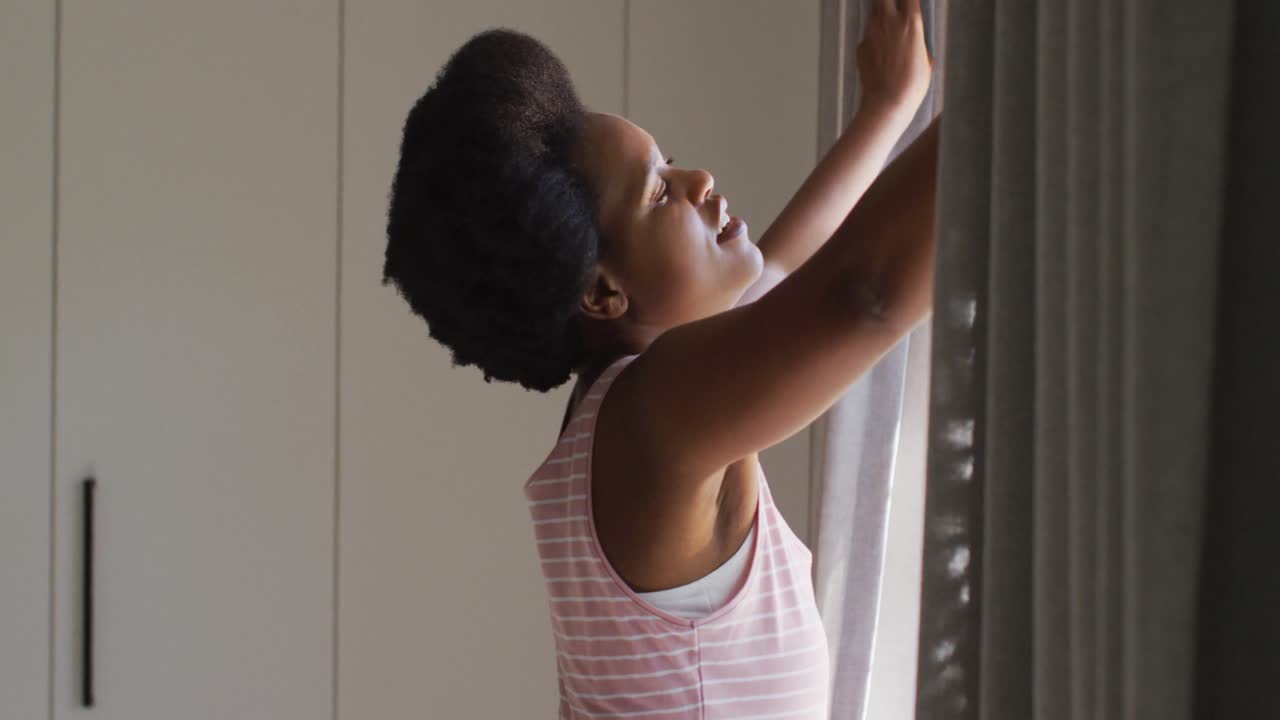Felice donna afroamericana che apre le tende in camera da letto.
