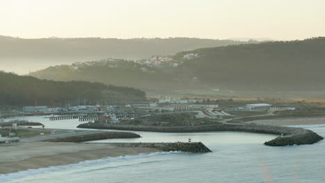 Praia-Da-Nazare-Mit-Porto-De-Abrigo-Und-Nebligen-Berg-Im-Hintergrund-Bei-Sonnenaufgang-In-Portugal