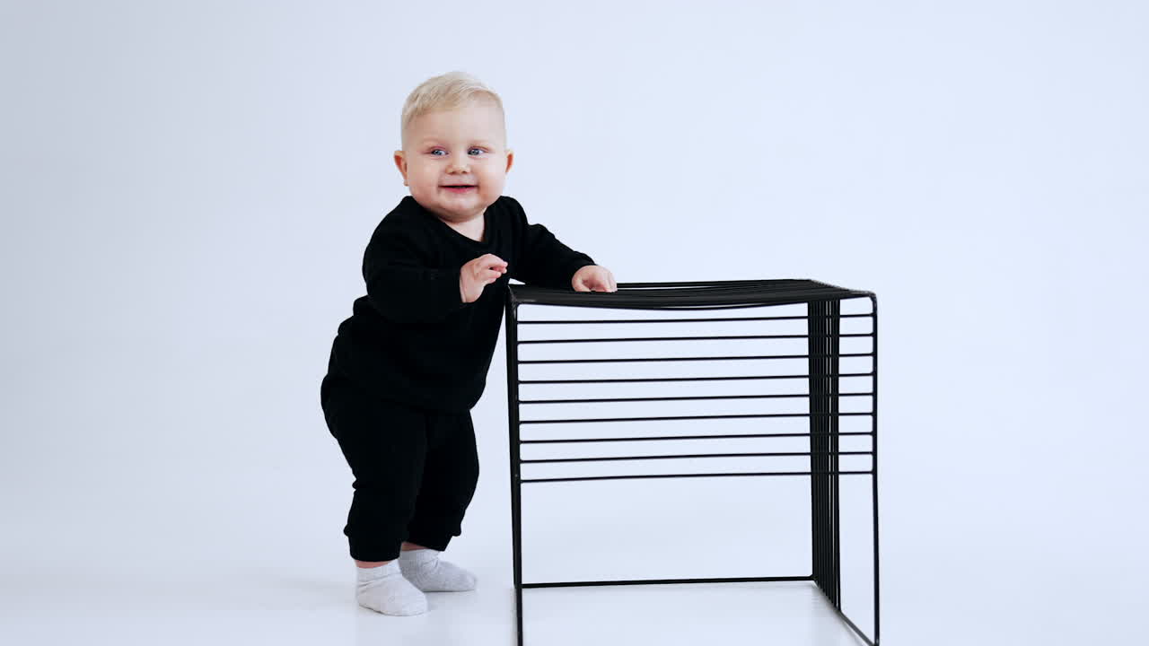 Adorable plump baby boy wearing black suit and white socks. Blond child stands holding by the cage. White backdrop.