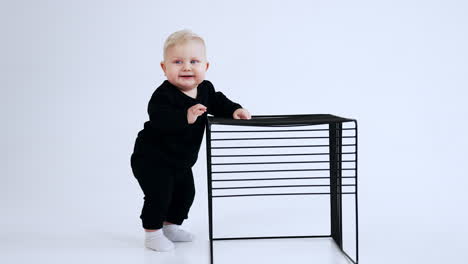 adorable plump baby boy wearing black suit and white socks. blond child stands holding by the cage. white backdrop.