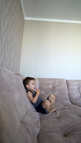 caucasian baby boy sitting on the big sofa. happy child throwing a toy to the camera. vertical video.