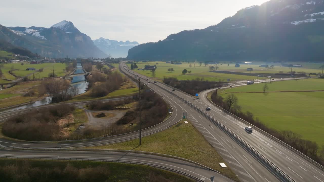Drone flight beside a highway with a circular on-ramp surrounded by open green fields and mountains. Clear daylight highlights vehicles moving through a scenic Swiss valley landscape