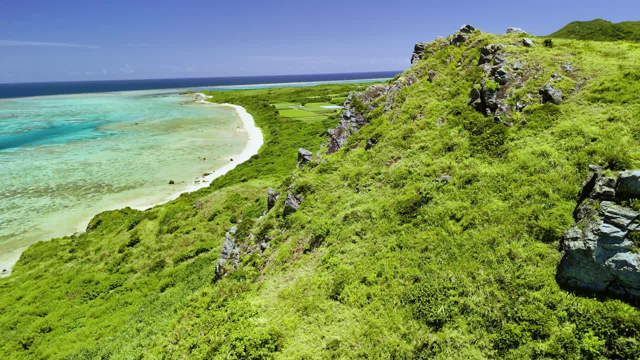Panoramica cinematografica con drone da lussureggianti scogliere rocciose e verdi a una spiaggia incontaminata di sabbia bianca e vaste barriere coralline turchesi sotto un cielo azzurro limpido sull'isola di Ishigaki, Giappone.