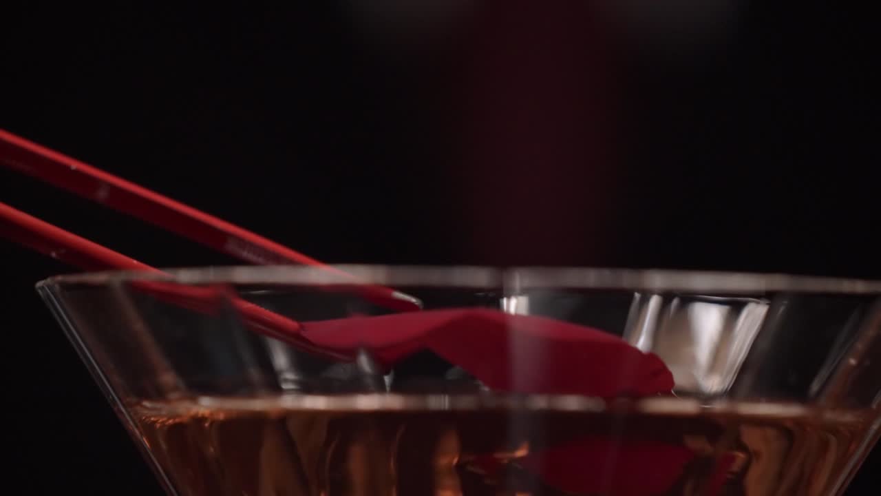 A bartender places a red rose petal on the top of the cocktail by using a red precision tongue.