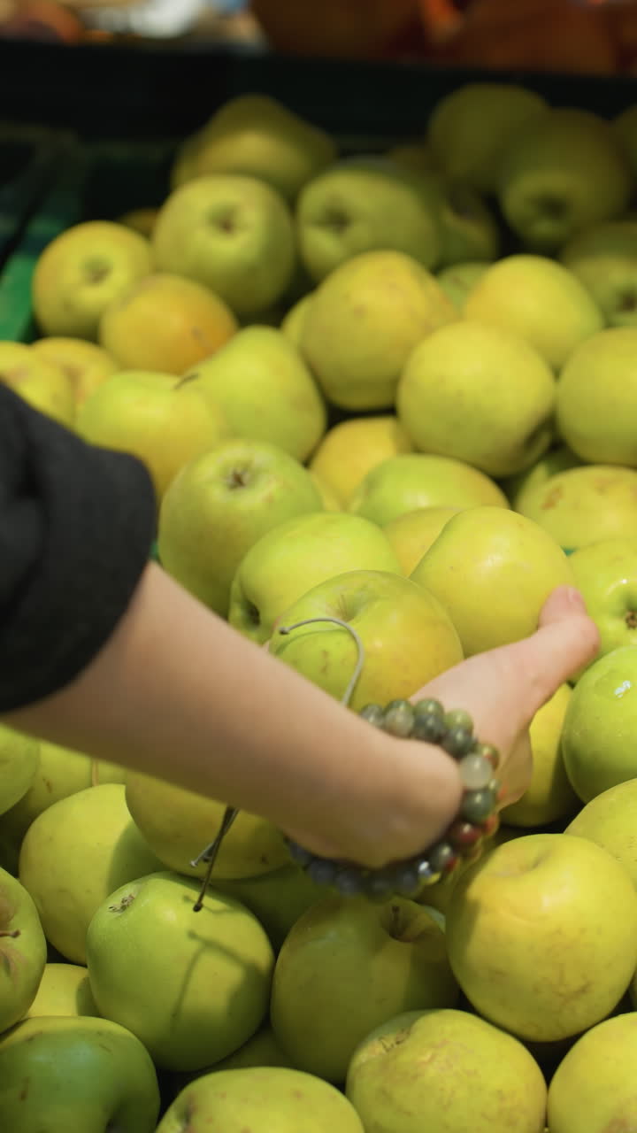 vista di una mano che indossa un cappotto nero e braccialetti di perle che raccoglie due mele da una cassa in un negozio di alimentari