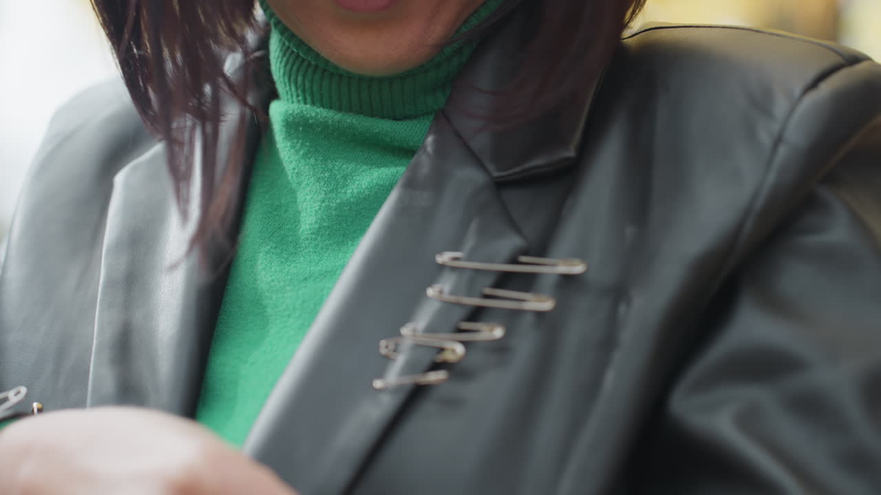 woman fastening safety pin on leather jacket, closeup showing row of metal pins used as brooch, green sweater visible, soft natural bokeh background, autumn light, candid handson styling, edgy DIY