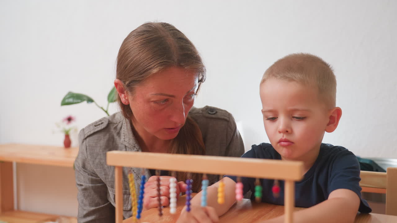 Primer plano de una madre guiando a un niño aburrido durante una lección con un ábaco de madera; el niño mueve las cuentas sin interés, con poco entusiasmo, entrenamiento de apoyo y un aula interior cálida.