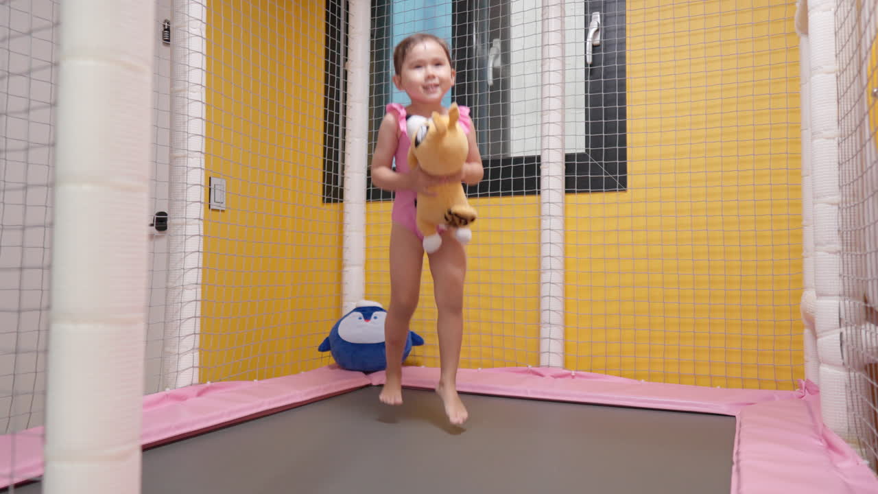 Excited Little 3-year-old Girl Jumping Barefoot on a Small Indoor Trampoline at Home Holding Favourite Plush Toy - slow motion