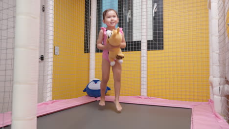 Excited-Little-3-year-old-Girl-Jumping-Barefoot-on-a-Small-Indoor-Trampoline-at-Home-Holding-Favourite-Plush-Toy---slow-motion