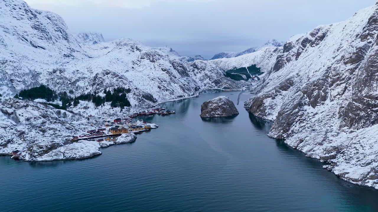 Eine atemberaubende filmische Luftaufnahme eines verschneiten Fjords und eines traditionellen Fischerdorfes auf den Lofoten, Norwegen. Zeigt die authentische nordische Winterlandschaft und das ruhige blaue Wasser.