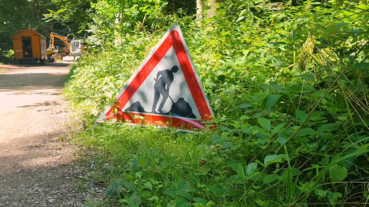 triangular warning sign in a Bern forest alerts pedestrians to forestry work, its vivid red contrasting with lush summer greenery in Switzerland
