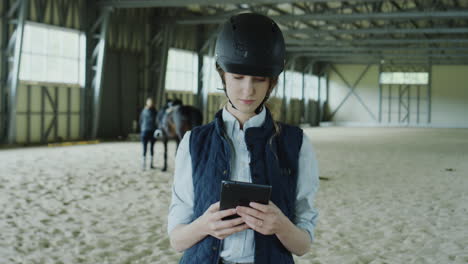 woman in equestrian gear holding a tablet in a riding arena with a horse