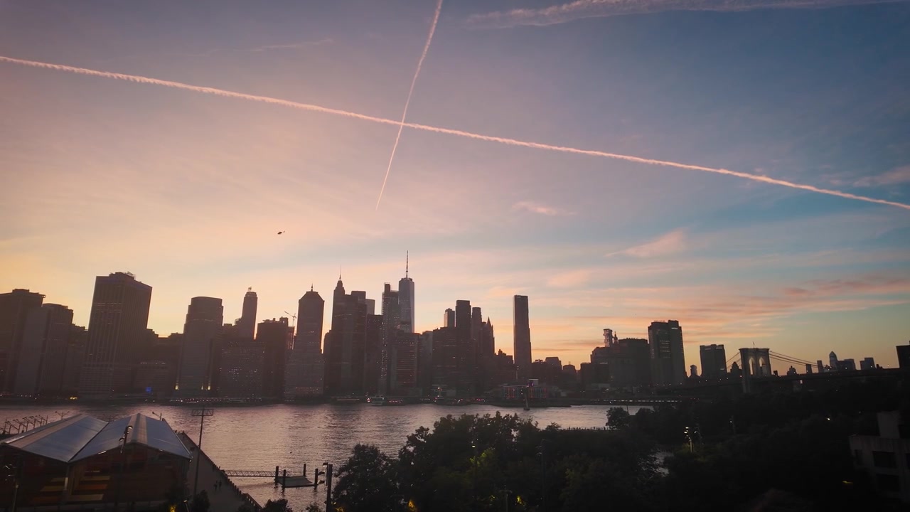 Gentle pan across the Lower Manhattan skyline at sunset from Brooklyn Heights as a helicopter crosses the colorful evening sky above New York City.