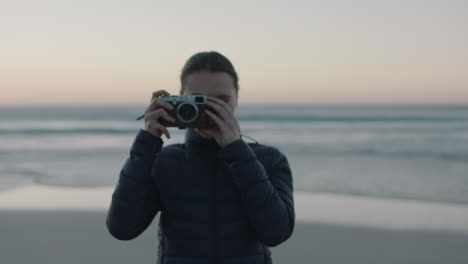 portrait of young confident woman taking photo using retro camera enjoying calm seaside beach at sunset