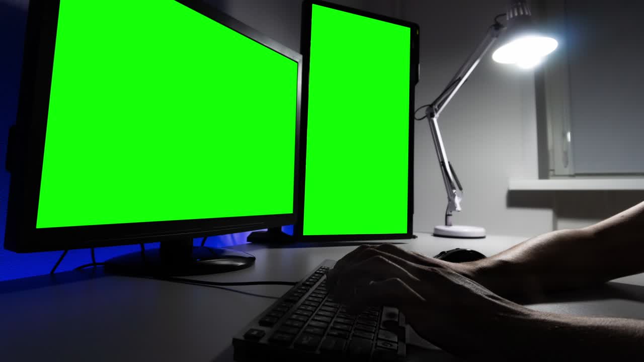 lapso de tiempo de las manos del hombre escribiendo en un teclado frente a dos monitores con canal alfa por la noche